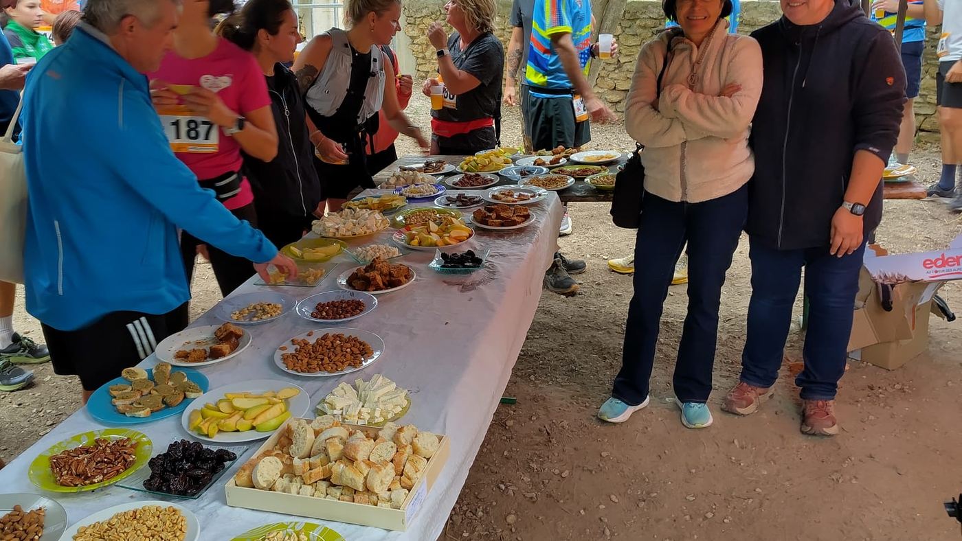 Deux bénévoles souriantes à côté de la table du ravitaillement, garnie de fruits secs, de pain, de fromage et de gâteaux maison.