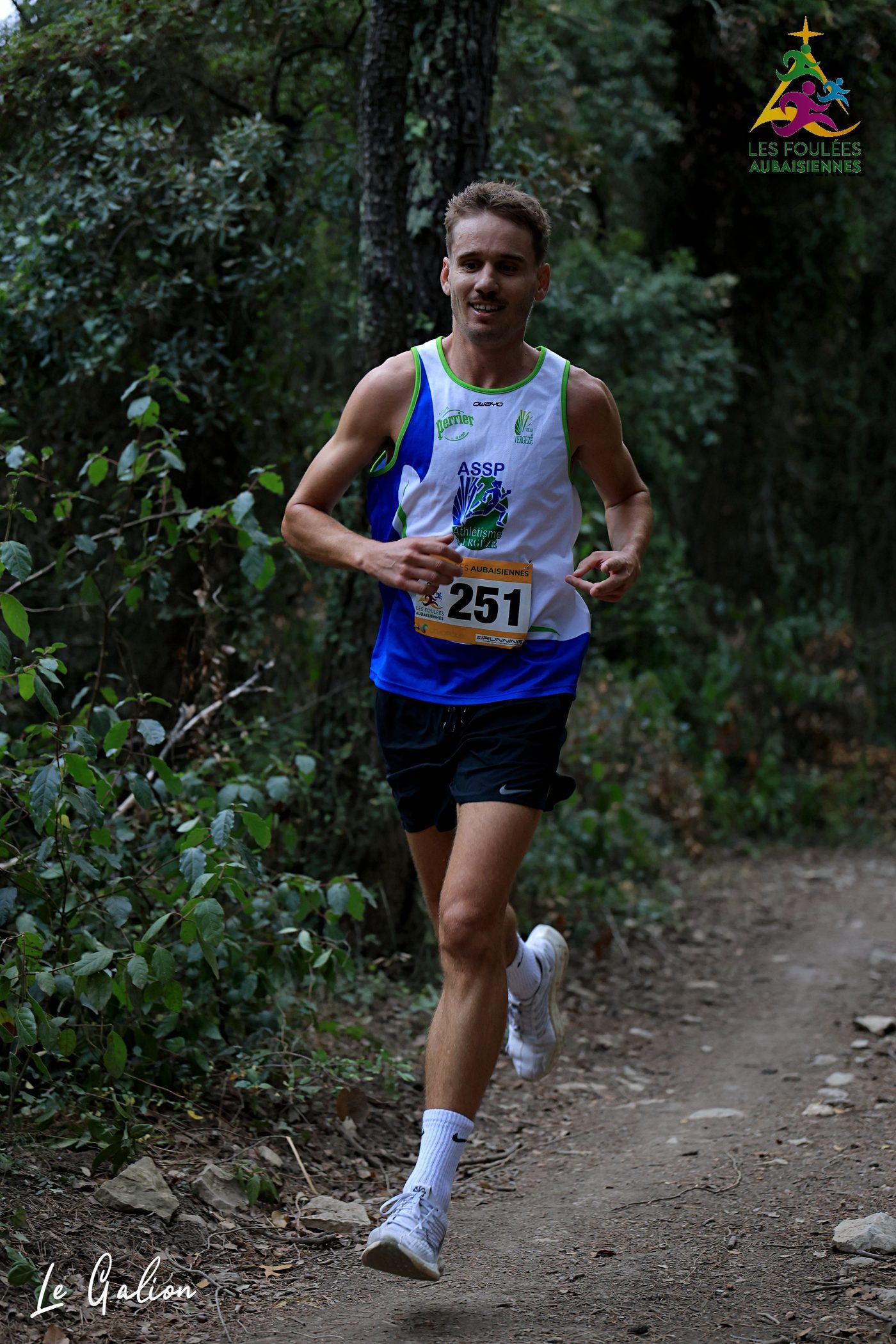 Un coureur en débardeur de club progresse sur un chemin de terre bordé de chênes verts.