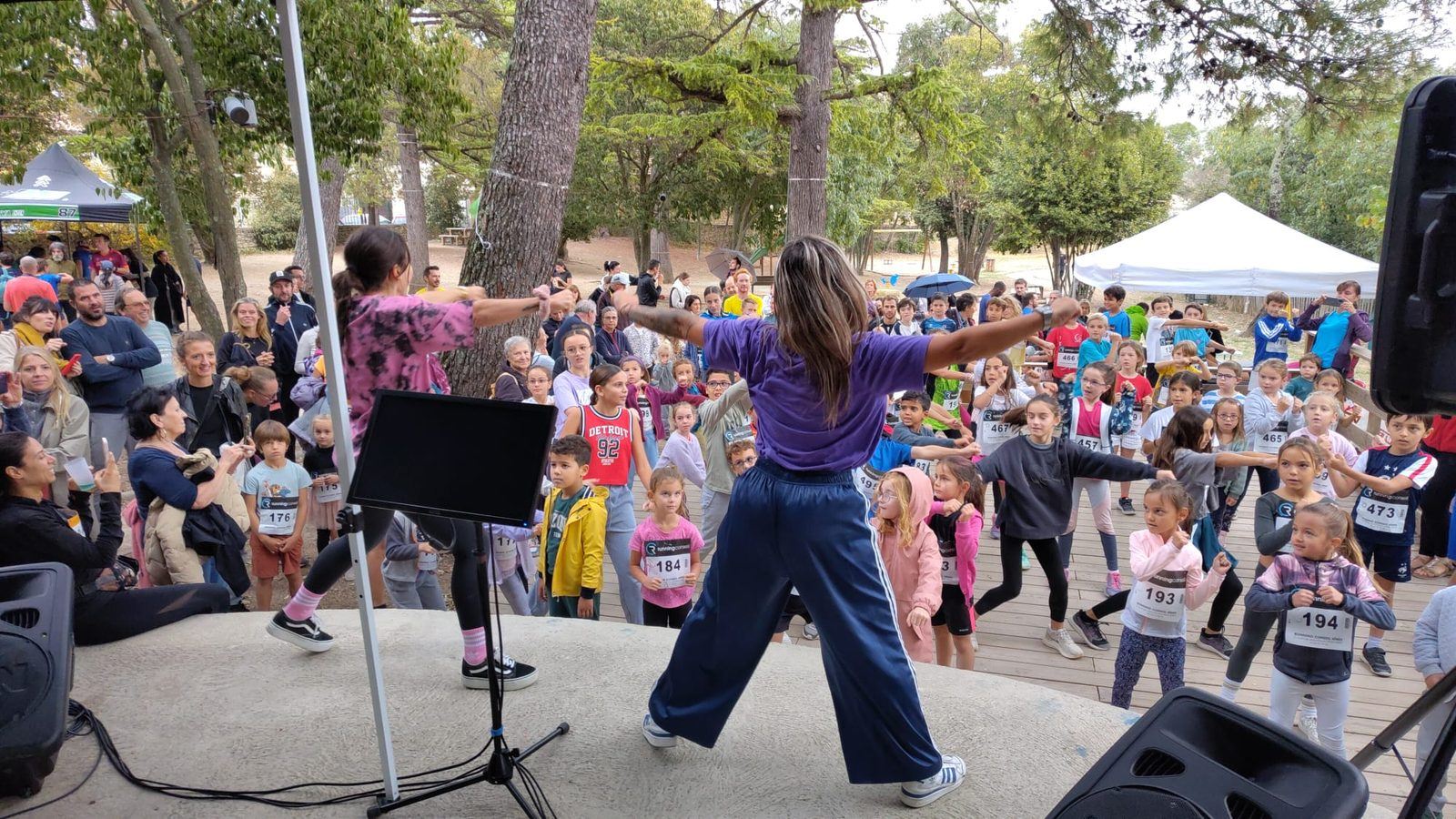 Des enfants avec leur dossard suivent l'échauffement dansé animé depuis la scène du parc.