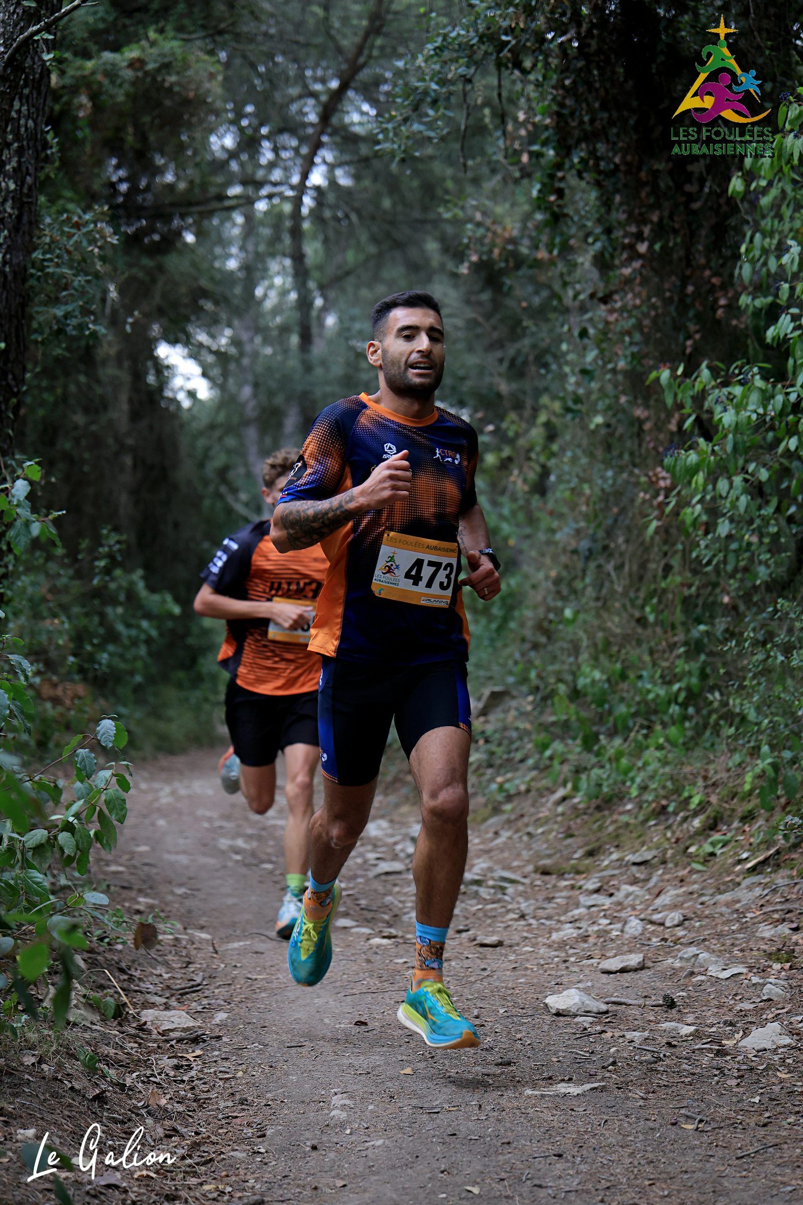 Un coureur en maillot de club sur un sentier de terre bordé de végétation, à l'ombre des chênes.